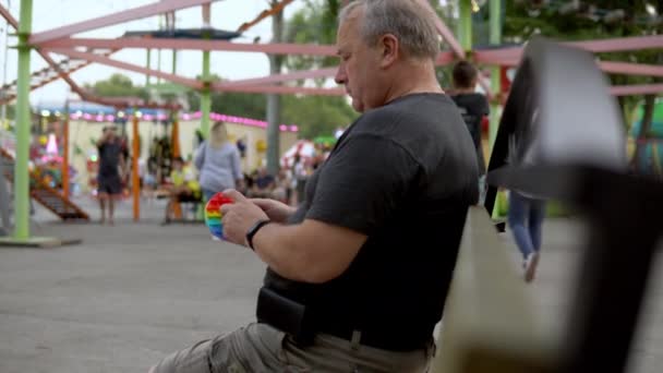 Ein Mann im Alter spielt in einem Popit im Hintergrund eines öffentlichen Parks. Die Person ist leidenschaftlich für Regenbogen-Anti-Stress. Silikonspielzeug für Kinder — Stockvideo