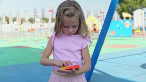 Дівчина в рожевій сукні грає епізод на тлі дитячого майданчика. Дитина захоплюється веселкою проти стресу. Дитяча силіконова іграшка — стокове відео