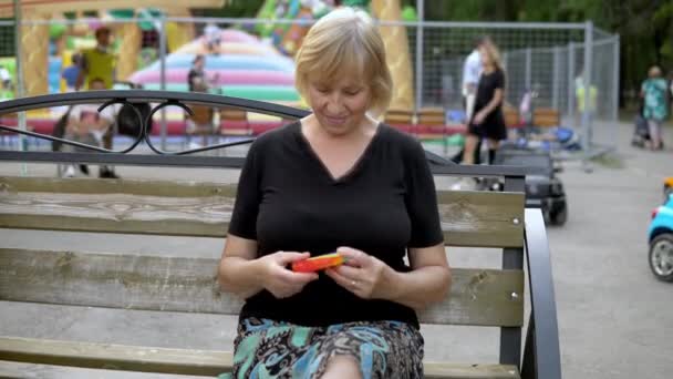 Eine betagte Frau spielt in einem Popit im Hintergrund eines öffentlichen Parks. Die Person ist leidenschaftlich für Regenbogen-Anti-Stress. Silikonspielzeug für Kinder — Stockvideo