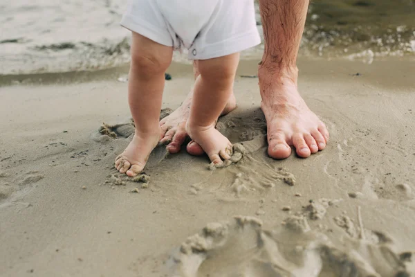 Bosé nohy otce a jeho malého syna v písku u vody. — Stock fotografie