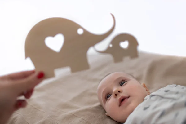 Early education concept. Portrait of the little toddler laying on beige blanket and looking to the wooden figure of elephant, which his mother showed to him — Stock Photo, Image