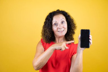 Orta yaşlı bir kadın, sarı arka planda dikilip gülümseyen ve telefonu işaret eden sıradan bir gömlek giyiyor.