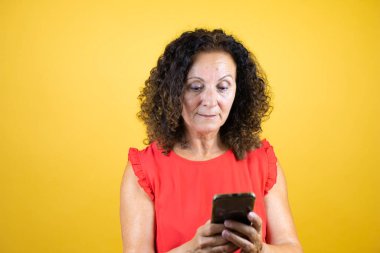 Orta yaşlı bir kadın, günlük gömleğini giymiş, izole edilmiş sarı arka planda duruyor ve telefonuyla mesajlaşıyor.