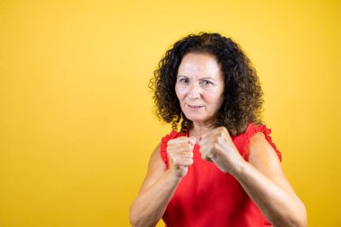 Orta yaşlı bir kadın sarı arka planda dikiliyor. Yumruklarını yumrukluyor, saldırıyor, saldırıyor, tehdit ediyor ve şiddet uyguluyor.