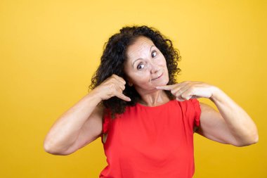 Orta yaşlı bir kadın, günlük gömleğini giymiş, sarı arka planda dikiliyor ve elleriyle bana jest yapıyor.