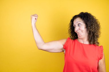 İzole edilmiş sarı arka planda duran sıradan tişörtlü orta yaşlı bir kadın gururla gülümseyen kol kaslarını gösteriyor.