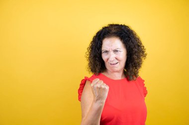 Orta yaşlı bir kadın, izole edilmiş sarı arka planda dikilmiş sinirli sinirli ve öfkeli bir şekilde yumruğunu kaldırırken öfkeyle bağırıyor. Öfke ve agresif kavram.