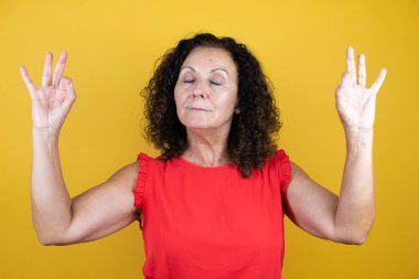 İzole edilmiş sarı arka plan üzerinde duran orta yaşlı kadın rahatla ve gözleri kapalı meditasyon yaparken parmaklarıyla gülümse.