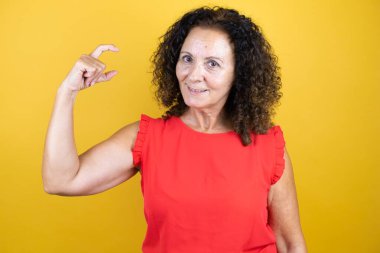 Orta yaşlı bir kadın, izole edilmiş sarı arka planda dikilmiş, gülümseyen, kendine güvenen, el hareketi yapan, parmaklarıyla işaret eden ve kameraya bakan sıradan bir gömlek giyiyor. Ölçü kavramı.