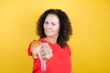 Orta yaşlı bir kadın, izole edilmiş sarı arka planda dikilen, kızgın yüzlü, negatif işaretli, başparmağı aşağıda olan, sıradan bir gömlek giyiyor.