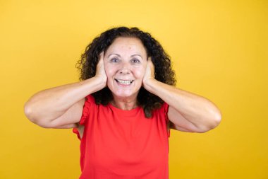 Orta yaşlı bir kadın, sarı arka planda dikilip duran sıradan bir tişört giyiyor. Deli ve korkmuş, elleri başında, şok geçirmekten korkan ve şaşıran biri.