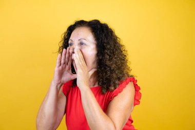 Orta yaşlı bir kadın, günlük gömleğini giymiş, sarı arka planda durmuş, elleri ağızda, bağırıyor ve bağırıyor.