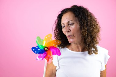 Orta yaşlı beyaz tişörtlü bir kadın izole edilmiş pembe arka planda duruyor elinde bir fırıldak tutuyor ve üflüyor.