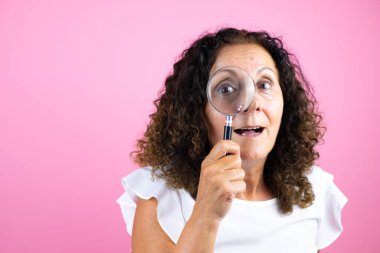 İzole edilmiş pembe arka planda duran beyaz gömlekli orta yaşlı bir kadın büyüteçten bakarken şaşırdı.