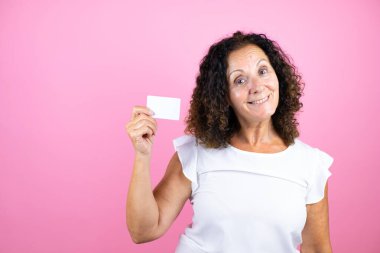 İzole edilmiş pembe arka planda duran beyaz tişörtlü orta yaşlı bir kadın gülümsüyor ve elinde beyaz bir kart tutuyor.