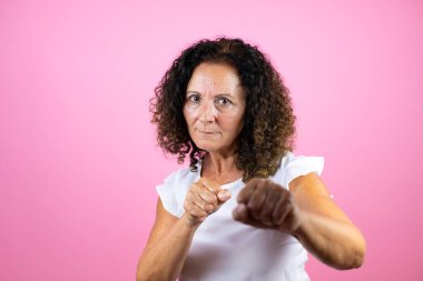 Orta yaşlı bir kadın, günlük beyaz gömlek giyiyor pembe arka planda duruyor dövüşmek için yumruk atıyor agresif ve öfkeli saldırı, tehdit ve şiddet.