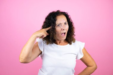 İzole edilmiş pembe arka planda dikilen beyaz gömlekli orta yaşlı bir kadın gülümseyerek ve parmakları kafasında bir fikri olduğunu düşünerek duruyor..