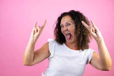 Orta yaşlı bir kadın, günlük beyaz gömlek giyiyor pembe arka plan üzerinde duruyor çılgın bir ifadeyle elleri havada rock sembolü yapıyor diye bağırıyor.