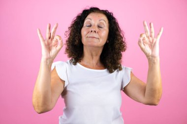 Orta yaşlı kadın, günlük beyaz gömlek giyiyor pembe arka planda dikiliyor rahatlıyor ve gözleri kapalı gülümsüyor parmaklarıyla meditasyon hareketi yapıyor.