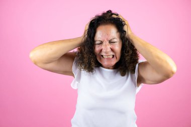 Orta yaşlı bir kadın pembe arka plan üzerinde dikilmiş baş ağrısı ve stresten muzdarip çünkü elleri başının üstünde acı ve migren var.