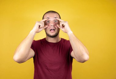 Rus adam, sarı tişörtlü, sarılı arka planda kırmızı tişört giyiyor. Gözlerini parmaklarıyla açmaya çalışıyor. Uykulu ve yorgun.