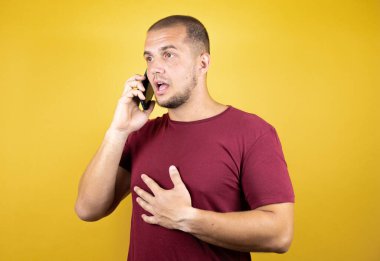 Rus adam sarı arkaplan üzerine kırmızı tişört giymiş. Sarı arka plan üzerinde akıllı telefon kullanarak sohbet ediyor. Şaşırmış bir yüzü var..