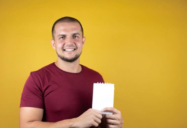 Rus adam, sarı tişörtlü, izole edilmiş arka planda gülümseyen ve elinde boş bir not defteri olan sıradan bir kırmızı tişört giyiyor.