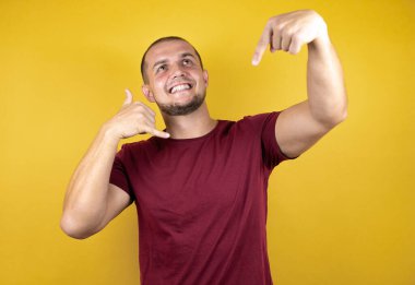 Rus adam, sarının üzerine kırmızı tişört giyip tebessüm ediyor. El ve parmaklarıyla telefon hareketi yapıyor. Telefonda konuşuyor ve elini işaret ediyor gibi.