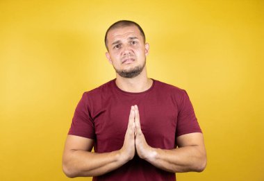 Rus adam, sarı arka plan üzerine kırmızı tişört giymiş, yalvarıyor ve dua ediyor. Yüzünde umut ifadesi var. Çok duygusal ve endişeli.