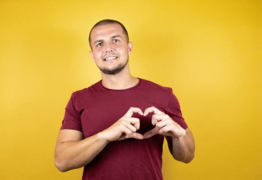 Rus adam, sarı izole arkaplan üzerine kırmızı tişört giymiş. Aşkla gülümsüyor. Elleriyle kalp sembolü ve şekli gösteriyor. Romantik konsept.