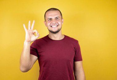 Rus adam sarı izole arkaplan üzerine kırmızı tişört giyip gülümsüyor ve el ve parmaklarıyla işaret veriyor. Başarılı ifade