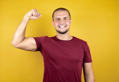 Rus adam sarı arka planda kırmızı tişört giyiyor. Kollarında gururla gülümseyen kaslar var.