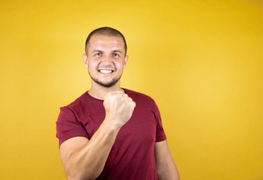 Sarı arka plan üzerine kırmızı tişört giyen Rus adam mutlu ve heyecanlı bir şekilde kollarını kaldırıp, gülümseyip başarı için haykırıyor..