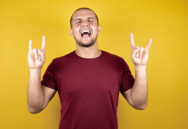 Rus adam, sarı izole arka plan üzerine kırmızı tişört giyip deli gibi bağırarak elleri havada rock sembolü yapıyor. Müzik yıldızı. Ağır konsept.