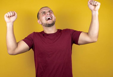 Sarı arka plan üzerine kırmızı tişört giyen Rus adam mutlu ve heyecanlı bir şekilde kollarını kaldırıp, gülümseyip başarı için bağırıyor. Kutlama konsepti.