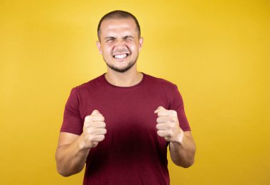 Sarı arka plan üzerine kırmızı tişört giyen Rus adam mutlu ve heyecanlı bir şekilde kollarını kaldırıp, gülümseyip başarı için bağırıyor. Kutlama konsepti.