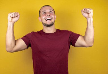 Sarı arka plan üzerine kırmızı tişört giyen Rus adam mutlu ve heyecanlı bir şekilde kollarını kaldırıp, gülümseyip başarı için bağırıyor. Kutlama konsepti.