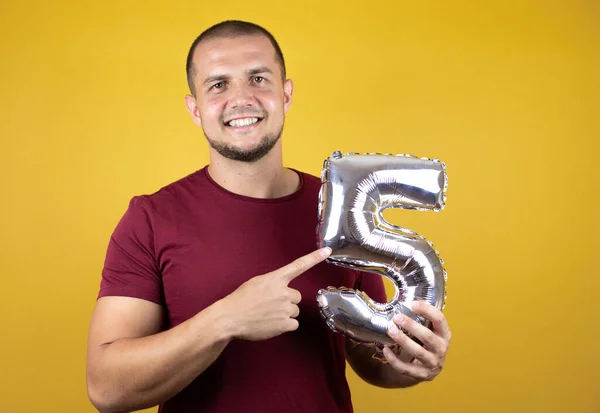 Russian man wearing basic red t-shirt over yellow insolated background smiling holding a number five. Aniversary