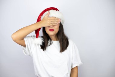 Beyaz arka planda Noel şapkası takan genç ve güzel bir kadın ciddi ve elleriyle gözlerini kapatıyor.