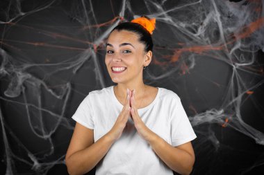 Young beautiful woman over black background with cobwebs and spiders hands together and fingers crossed smiling relaxed and cheerful. Success and optimistic. clipart