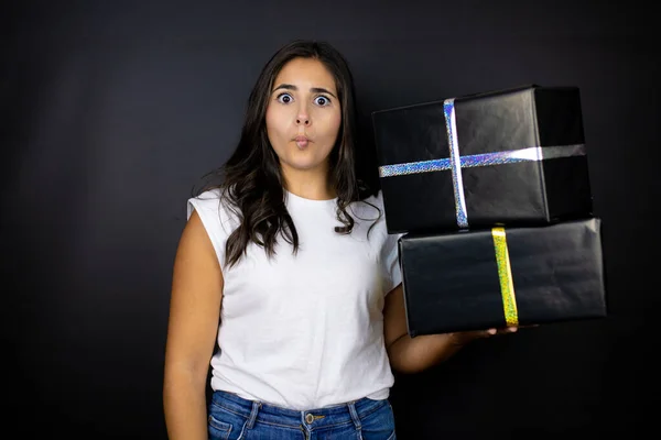 Young beautiful woman holding gifts over isolated black background making fish face with lips, crazy and comical gesture.