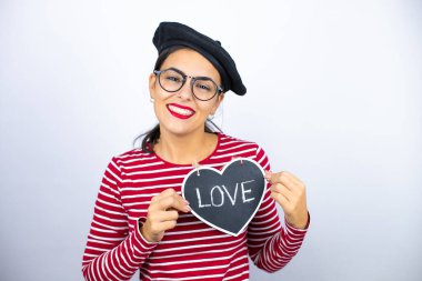 Fransız beresi takmış, beyaz arka planda gözlüklü güzel esmer kadın gülümsüyor ve elinde aşk mesajıyla kalp tahtası tutuyor.