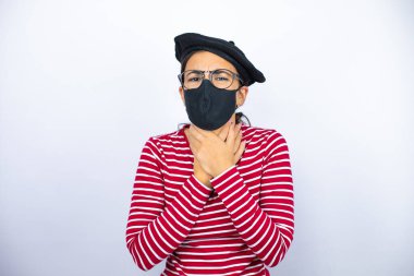 Fransız beresi takan, beyaz arka planda gözlük takan, maske takan, boynu ağrıyan, grip için boğaz ağrıyan ve enfeksiyon kapmış genç, güzel bir esmer kadın.