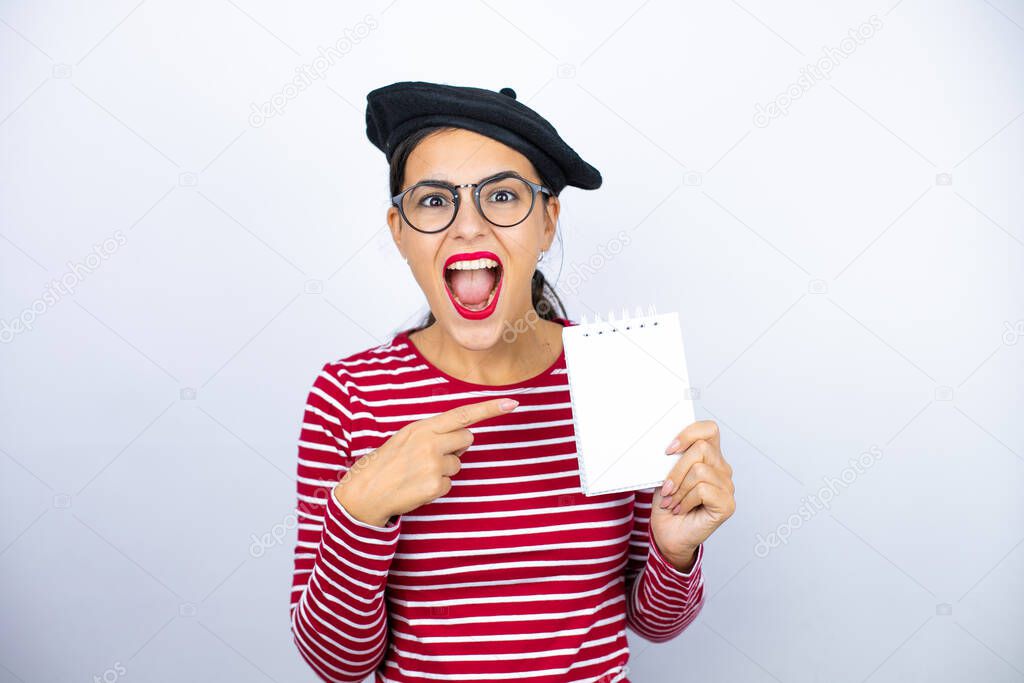Young beautiful brunette woman wearing french beret and glasses over white background smiling, surprised and pointing blank notebook in her hand