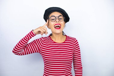 Fransız beresi takmış, beyaz arka planda gözlük takmış, gülümseyen ve parmakları kafasında bir fikri olduğunu düşünen genç ve güzel bir esmer kadın..