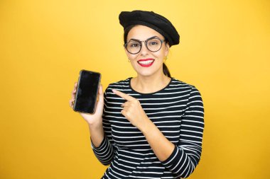 Fransız beresi takmış, sarı arka planda gözlüklü güzel esmer kadın gülümsüyor ve telefonu işaret ediyor.