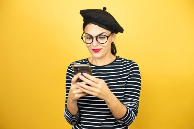 Fransız beresi takmış, sarı arka planda gözlüklü güzel esmer kadın telefonuyla mesajlaşıyor.