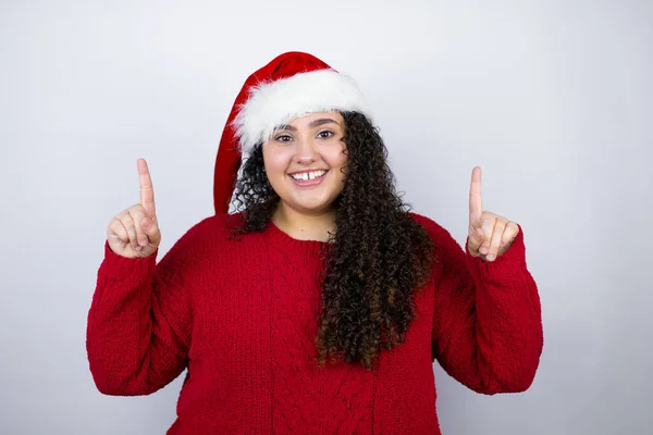 年轻美丽的女人 头戴圣诞帽 头戴白色背景 面带微笑 看着相机 用手指和胳膊指指着 — 图库照片