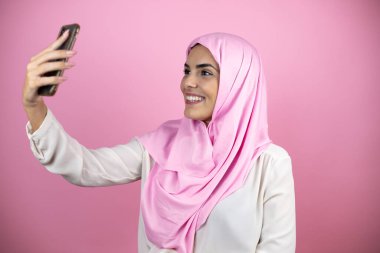 Genç, güzel bir Arap kadın, izole edilmiş pembe arka planda İslami başörtüsü takıyor telefonuyla selfie çekiyor.
