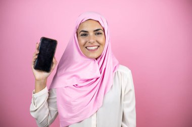 Genç, güzel bir Arap kadın, izole edilmiş pembe arka planda, tebessüm ederek ve telefonu işaret ederek İslami başörtüsü takıyor.